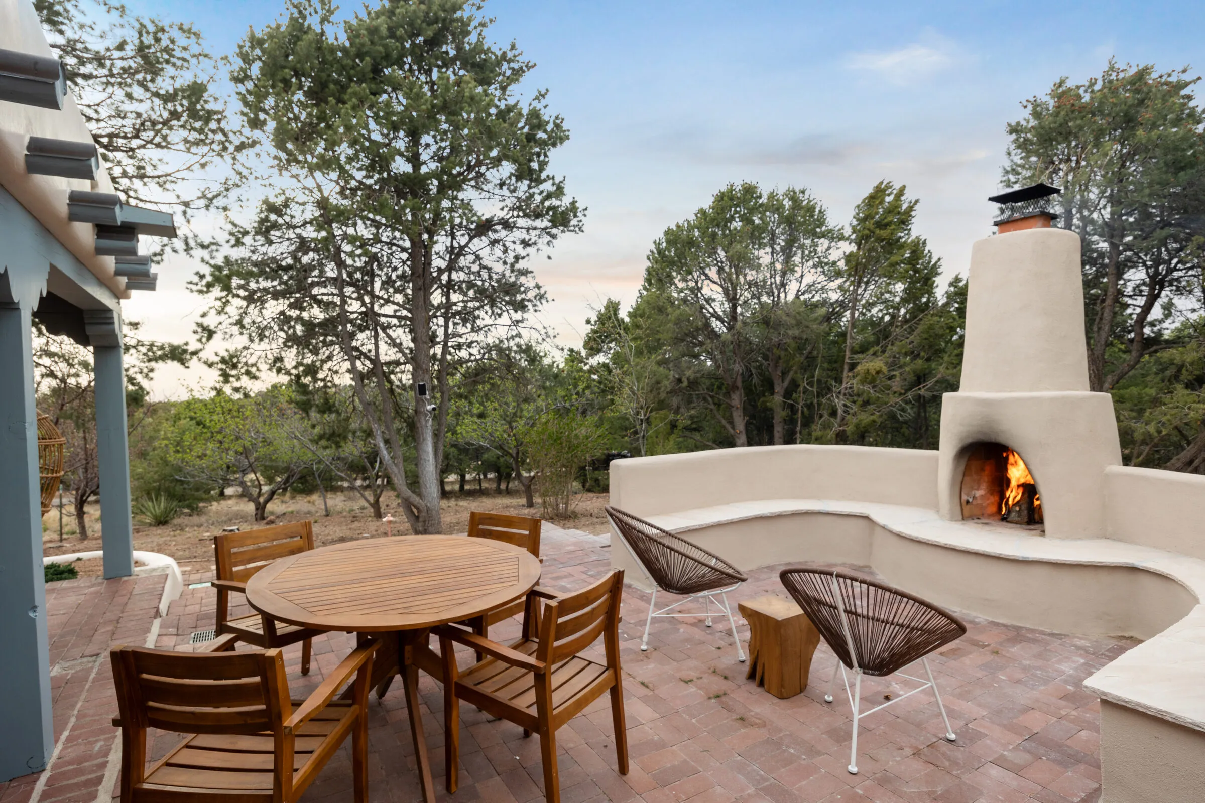 The outdoor kiva fireplace at Casa de Ice, lit at dusk — curved bench, teak table, and chairs under the trees