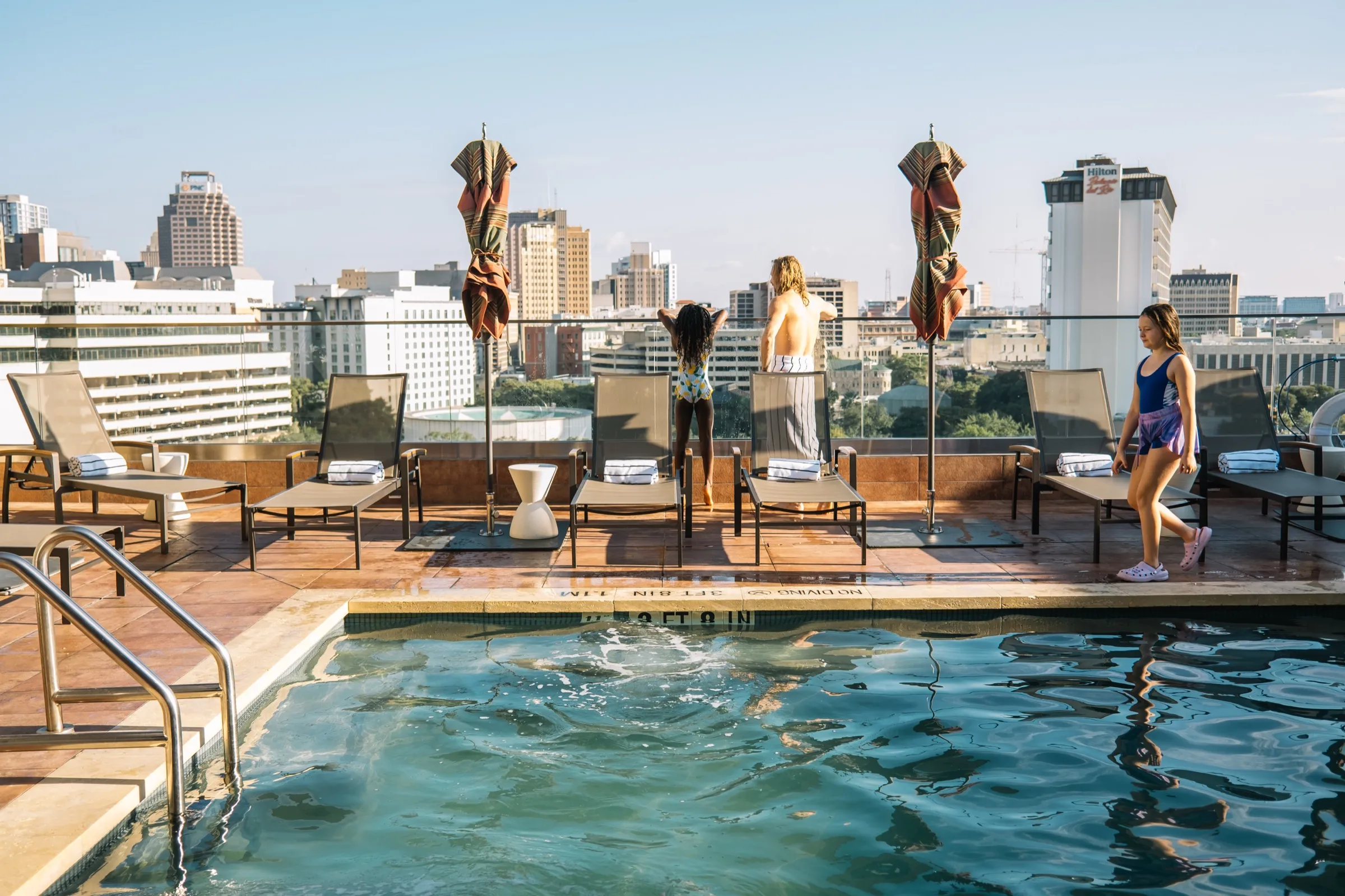 Melissa and Justice at the glass rail of the Kimpton Santo rooftop pool with the San Antonio skyline behind them, Rosie walking past the loungers